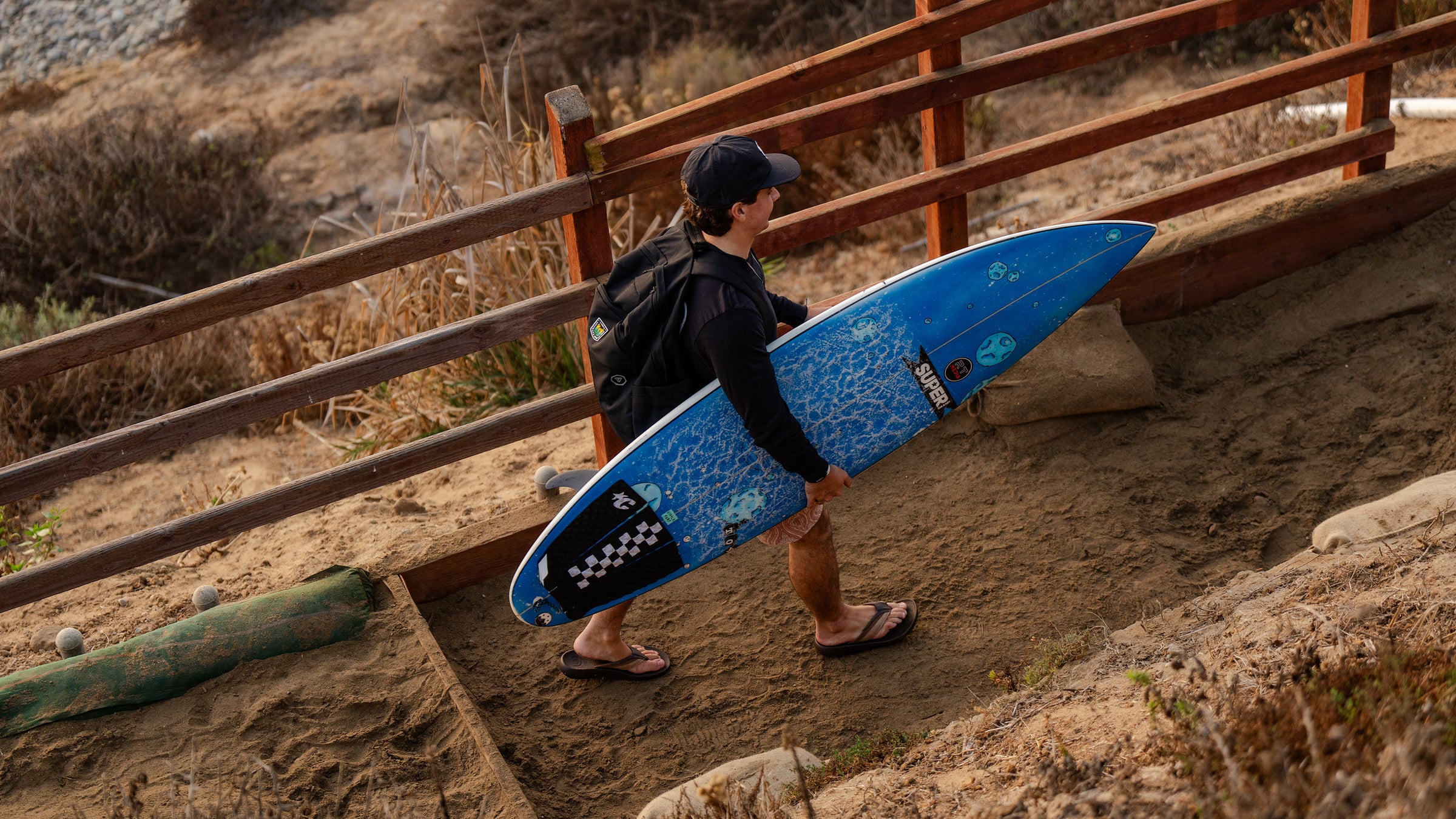 Man carrying blue surfboard along sandy path beside a wooden fence