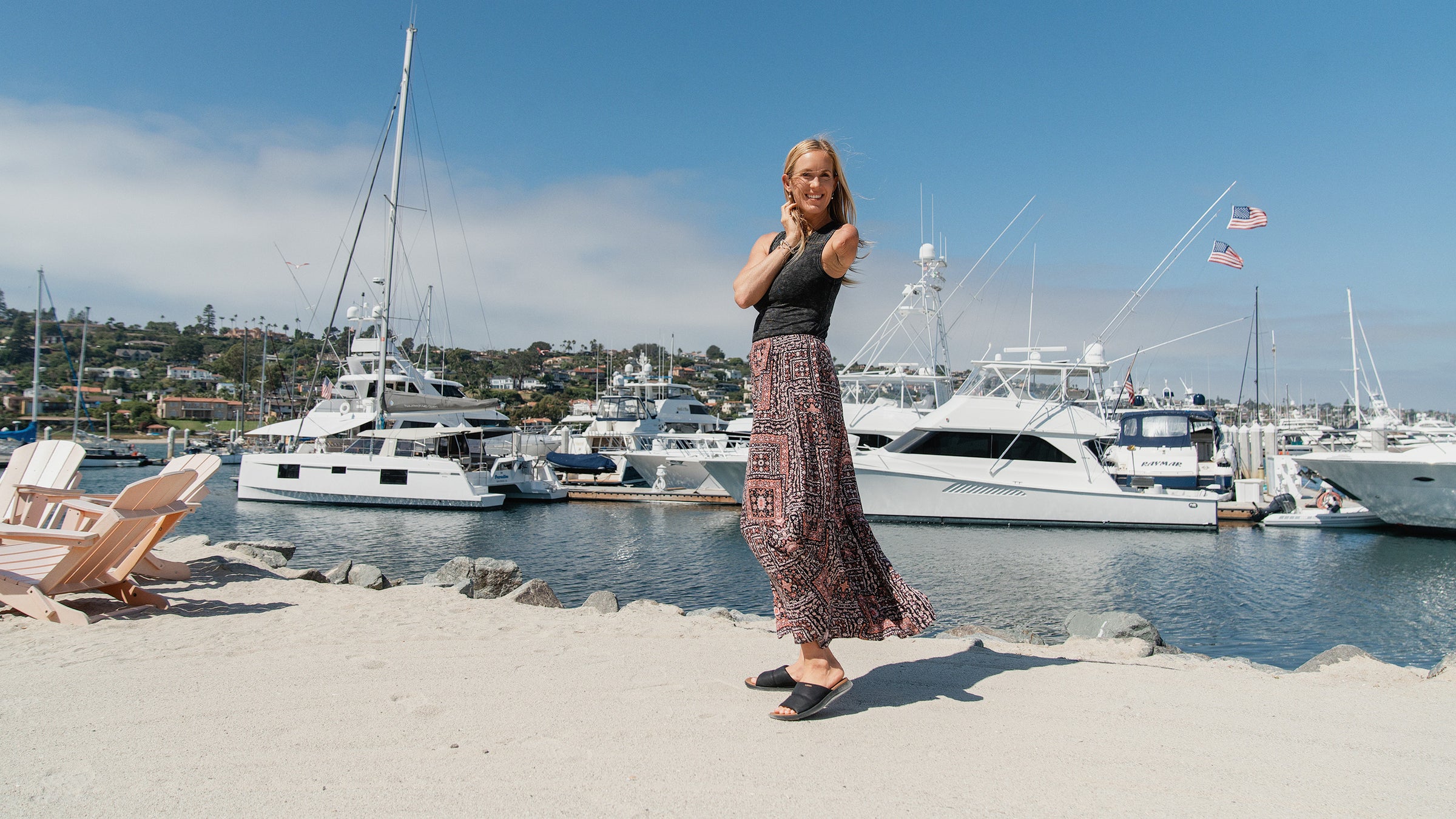 Woman standing on a sandy beach near a marina with yachts and American flags in the background
