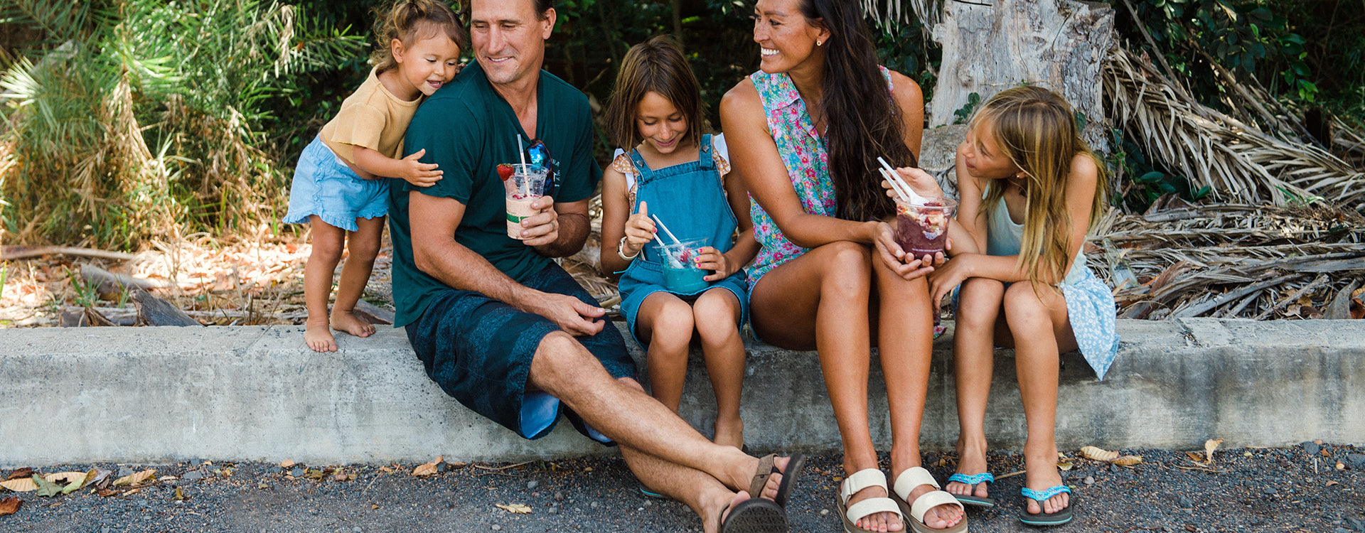 Family of five sitting on a concrete curb enjoying smoothies and desserts outdoors.
