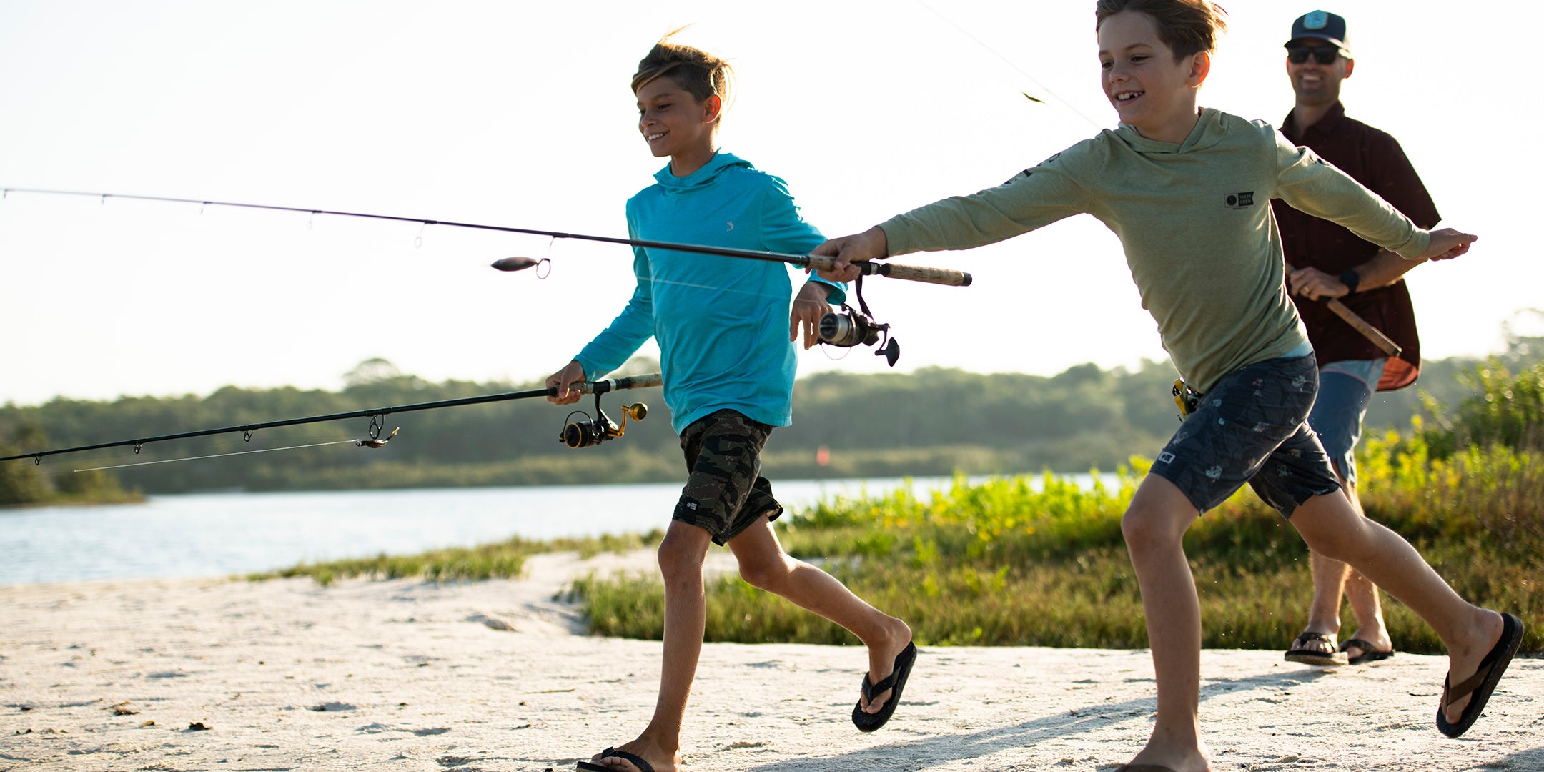 two boys running along a sandy riverbank with fishing rods