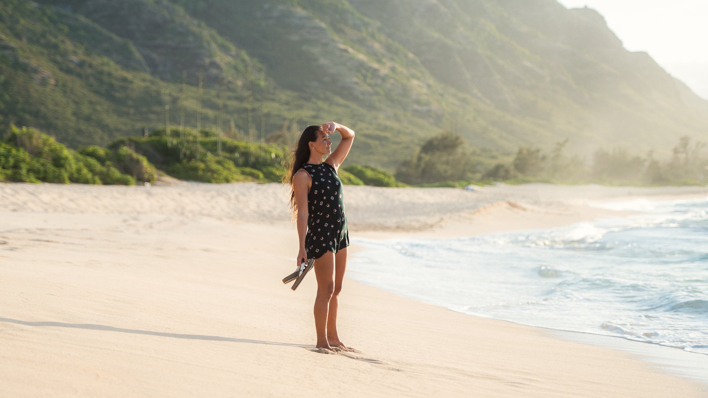 Corinne Gold walking a beach with sandals in hand, looking out over the ocean.