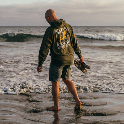 CJ Hobgood walking in the water at a sandy shore with Cobian Anchor tan sandals in hand #color_tan