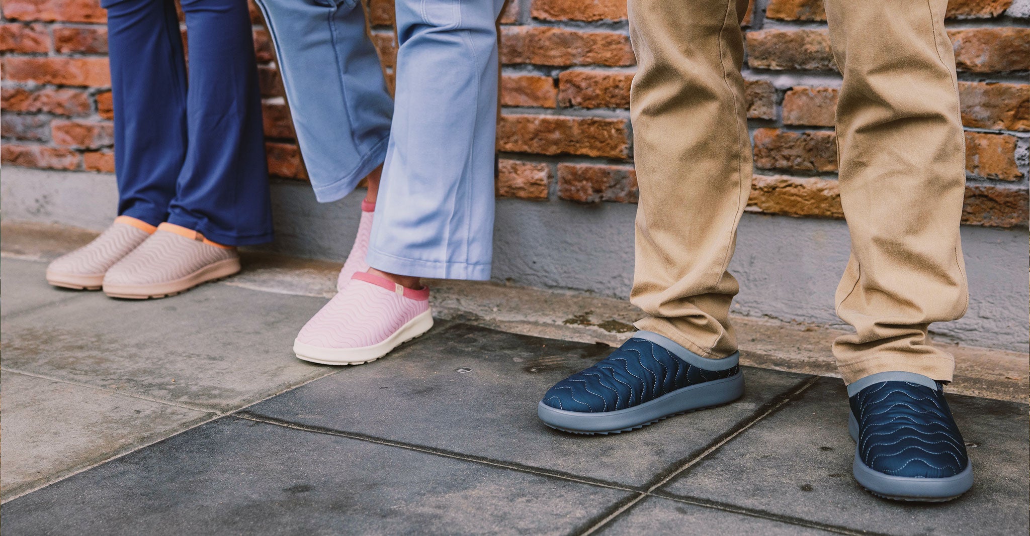 Three pairs of feet wearing different colored shoes on a stone floor with a brick wall background.