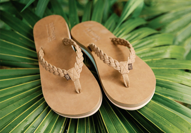 Tan braided-strap flip flops resting on green tropical leaves