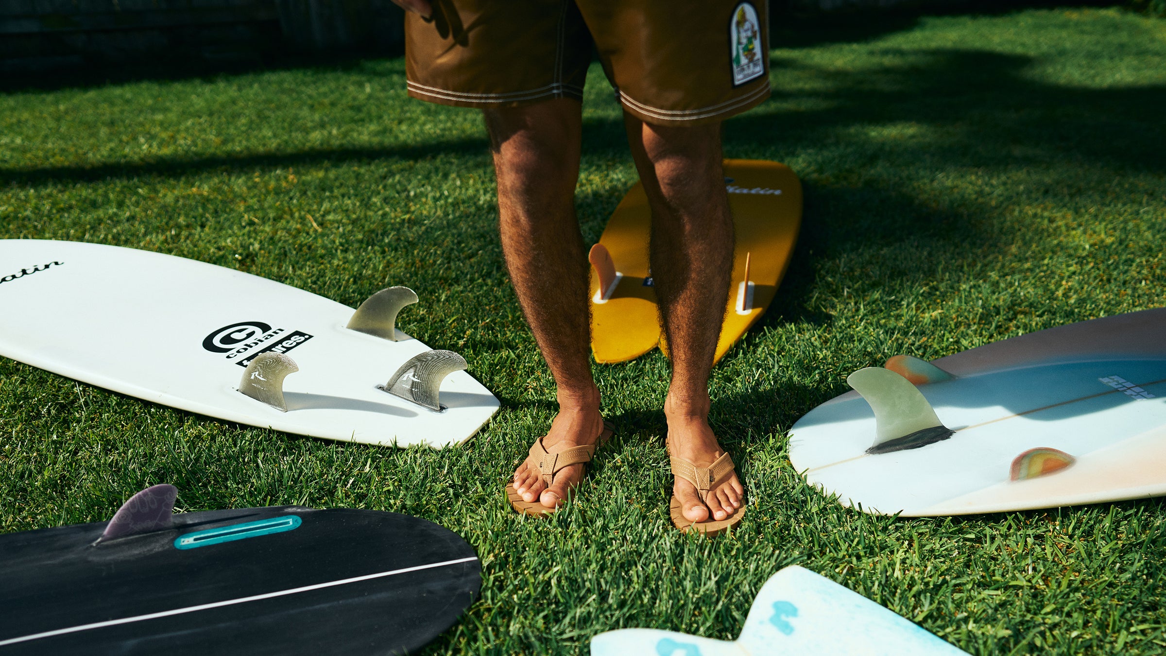 Model wearing Las Olas 2 is standing in grass surrounded by surfboards #color_tan