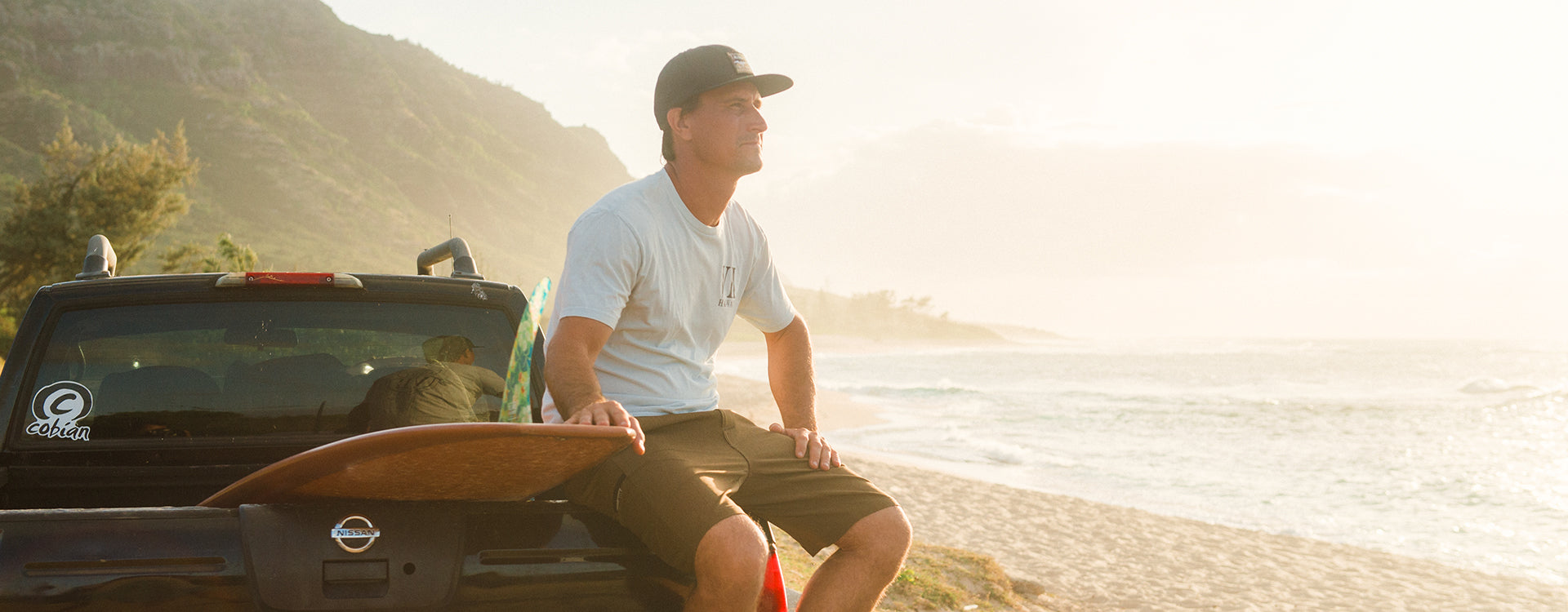 Aaron Gold sitting on a tailgate with his surfboard  looking over the ocean.